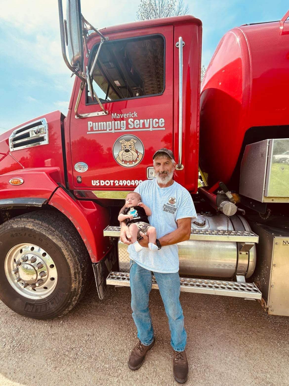 Man standing beside a red plumbing service truck, holding a baby in front of the cab.