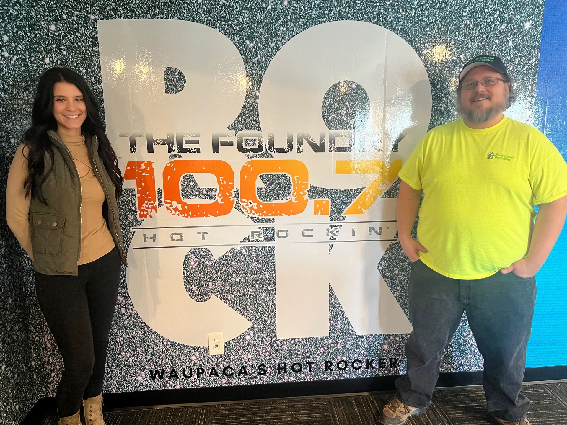 Two people standing in front of a glittery radio station logo wall, one in black and tan, one in neon yellow.