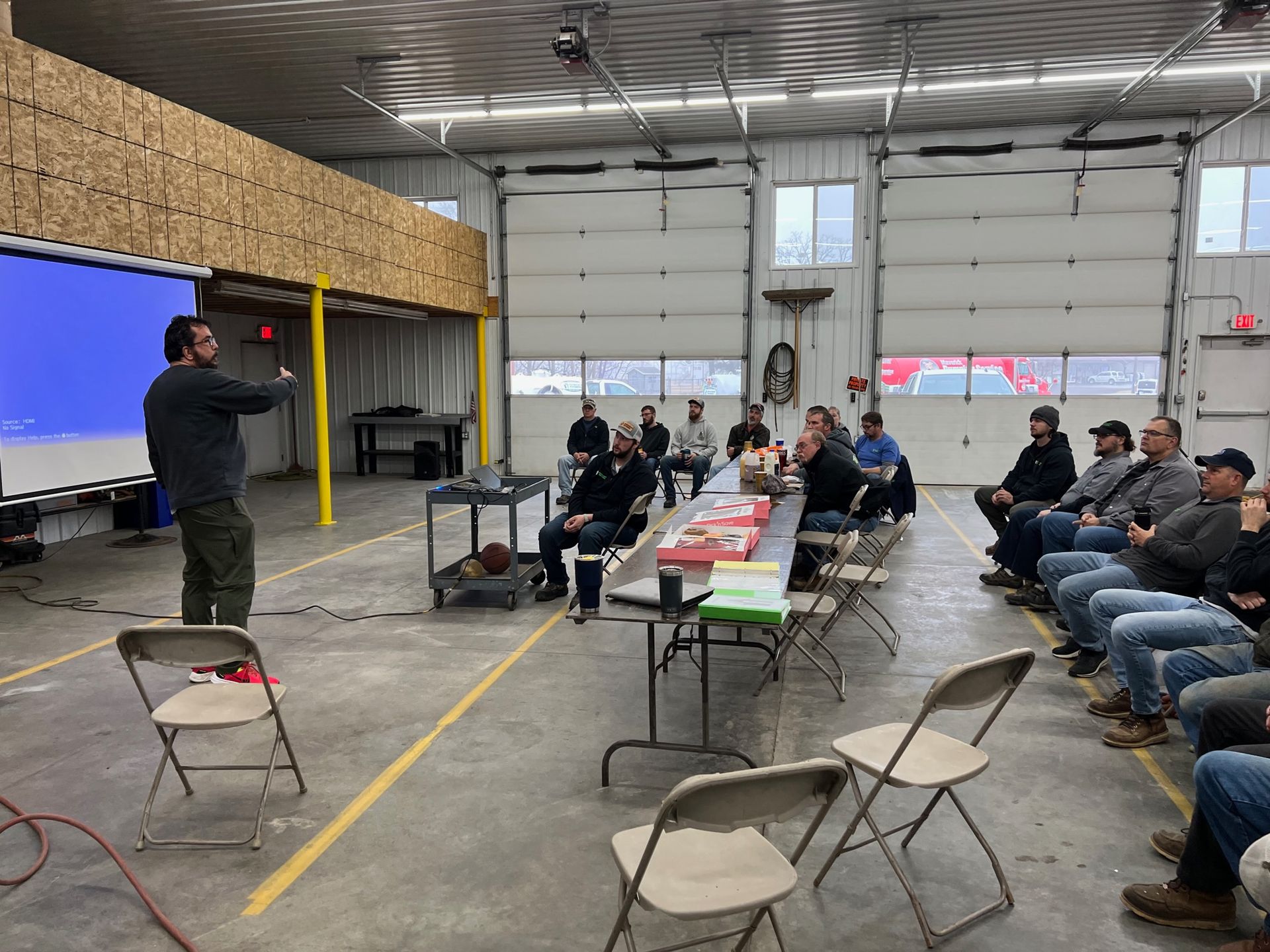 Speaker addressing seated group in warehouse, with folding chairs and tables arranged for a presentation