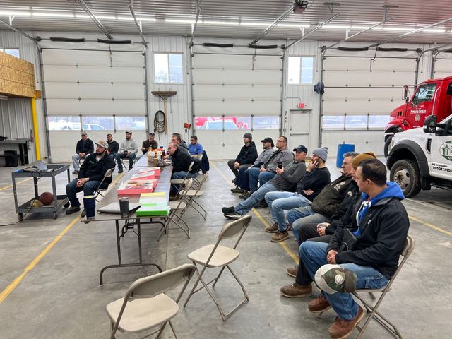 People seated in a garage workshop around folding tables, listening during a meeting with a red truck nearby.