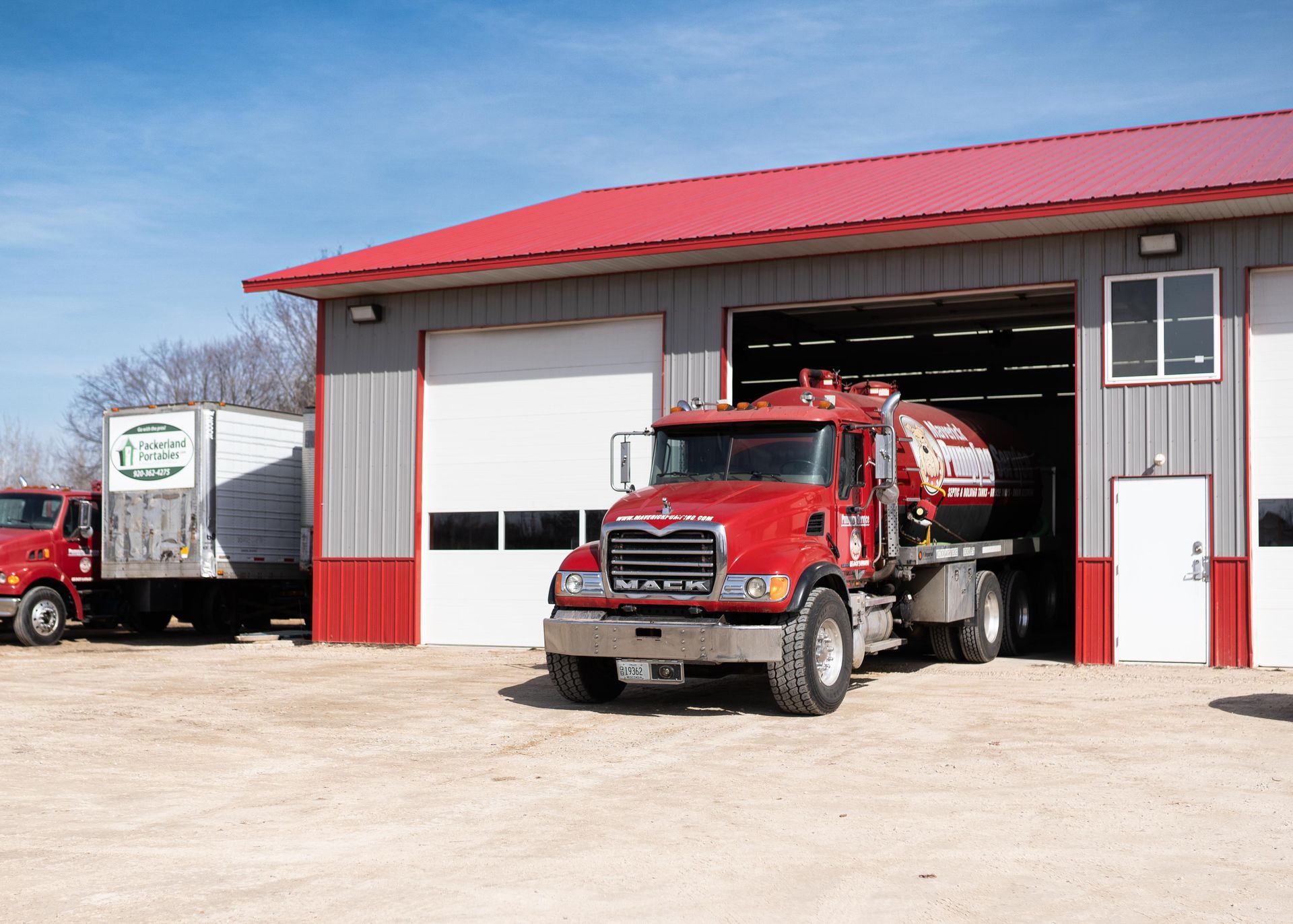 Red tanker truck outside a fire station with open garage bay and a white trailer nearby