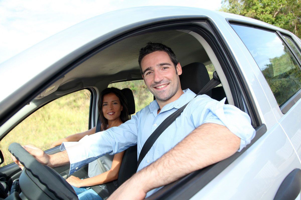 Man smiling, driving a car with a passenger. Both are wearing seatbelts.