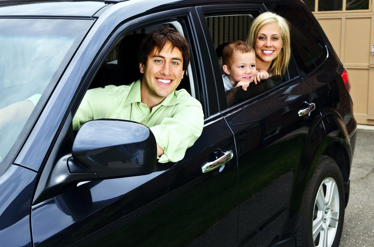 Family of three smiling from inside a black car: Father driving, mother and child in the back.