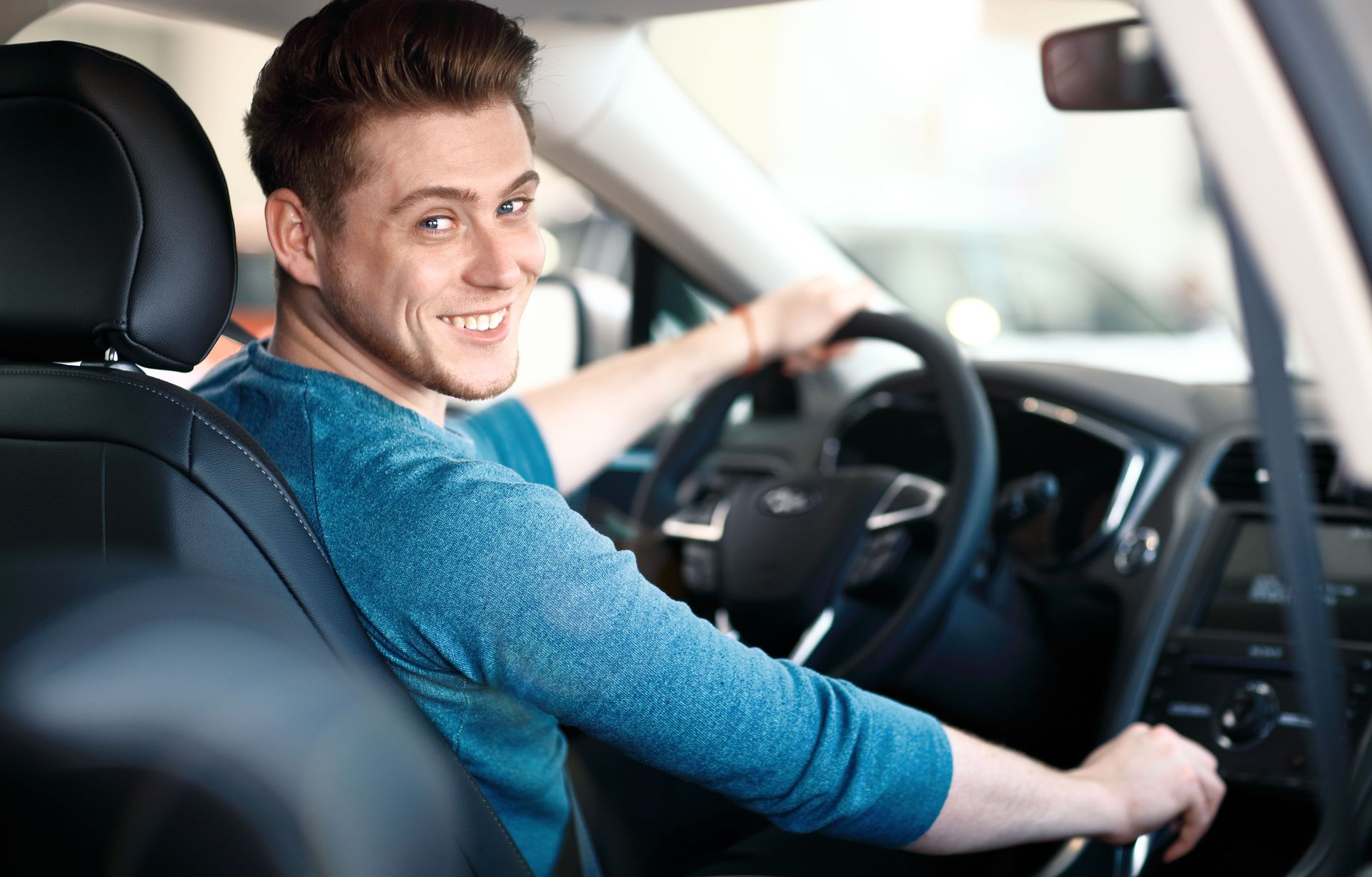 Man sitting in a car, smiling while holding the steering wheel.