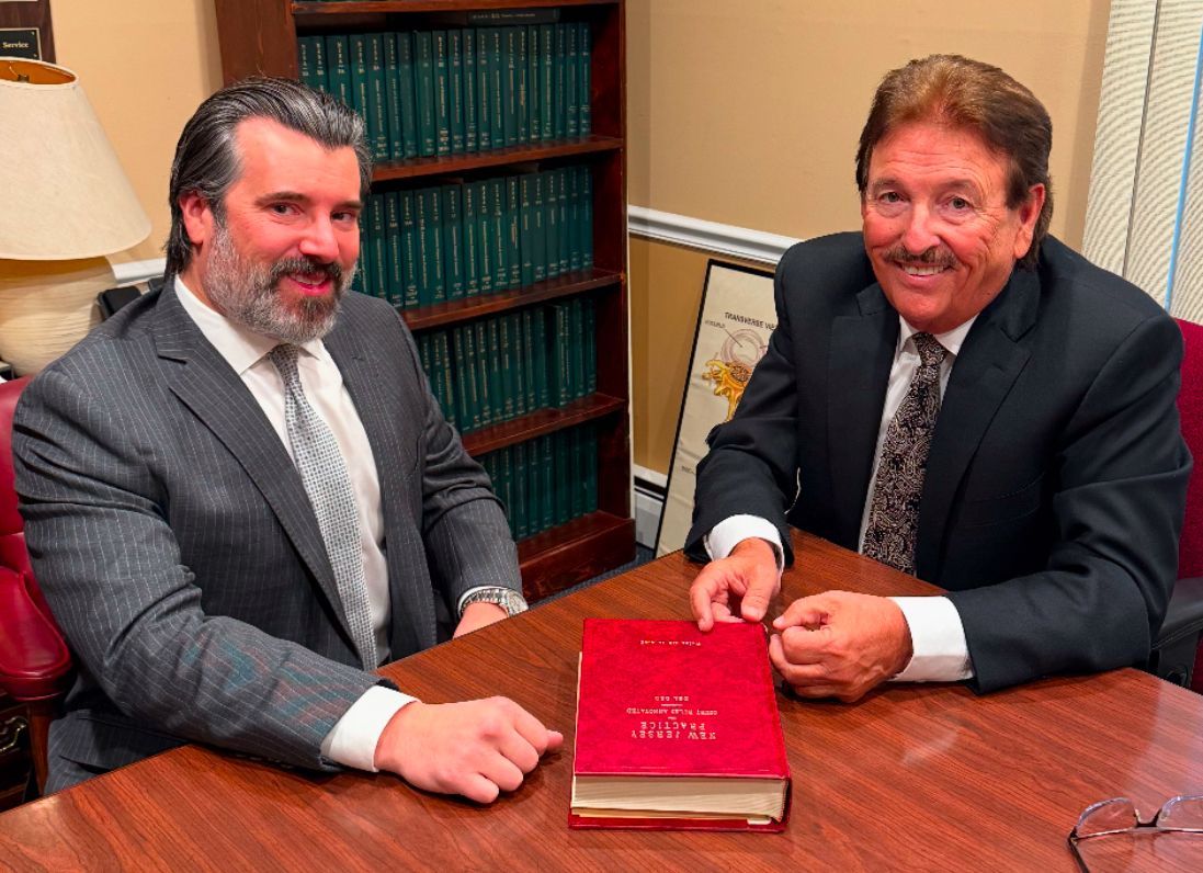 Two men seated at a table, holding a red book. They are in a room with a bookshelf.