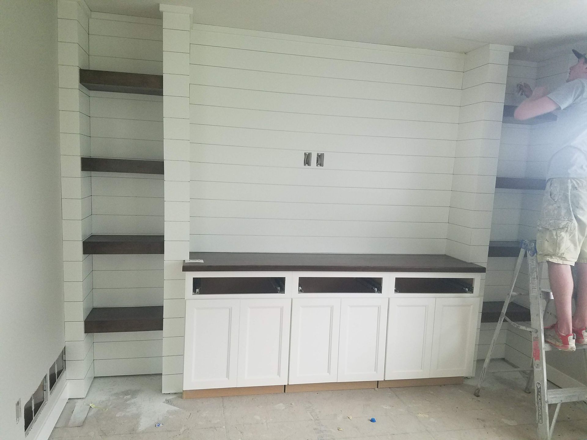 A person is standing on a ladder in a room with shelves and cabinets.