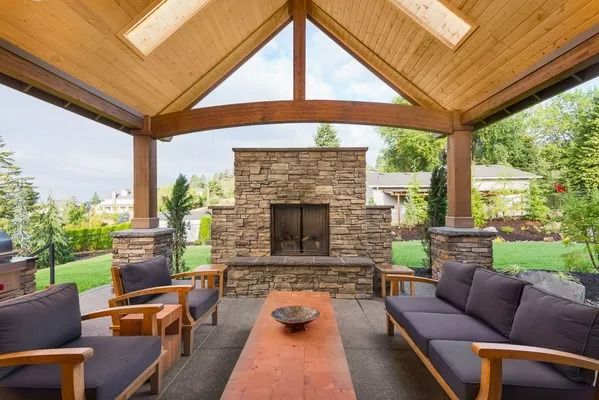 Covered patio seating area with stone fireplace, wood ceiling, and outdoor sofas вокруг a coffee table