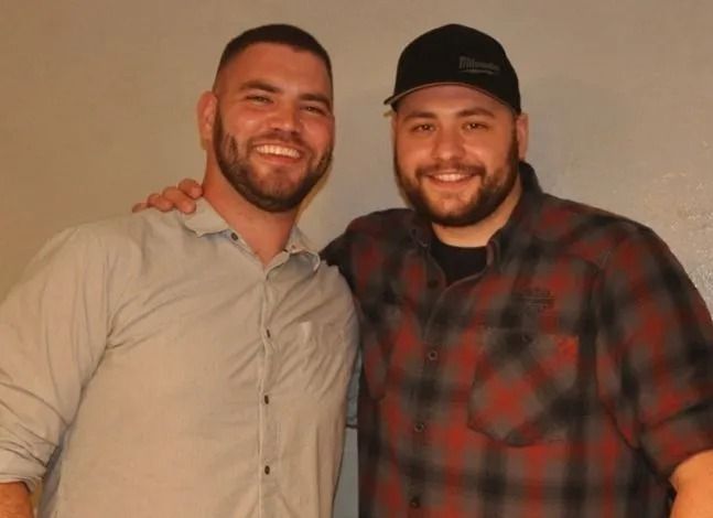 Two smiling men standing close together, one in a light shirt and one in a red plaid shirt with a black cap.