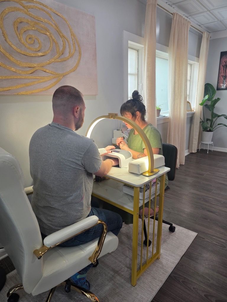 Two people at a manicure table in a bright salon, one painting the other's nails.