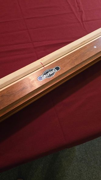 A beige pool table with dark wood trim in a game room, beside a red-felt table and windows.