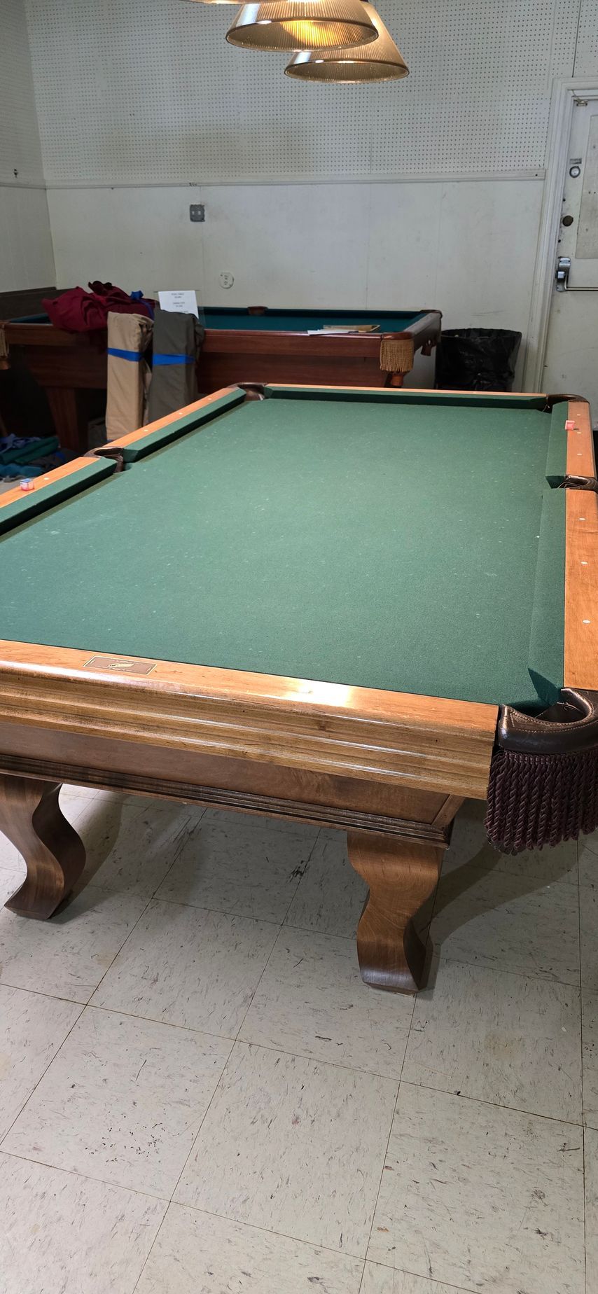 Green-felt pool table in a small game room with overhead lights and a wooden bench in the background