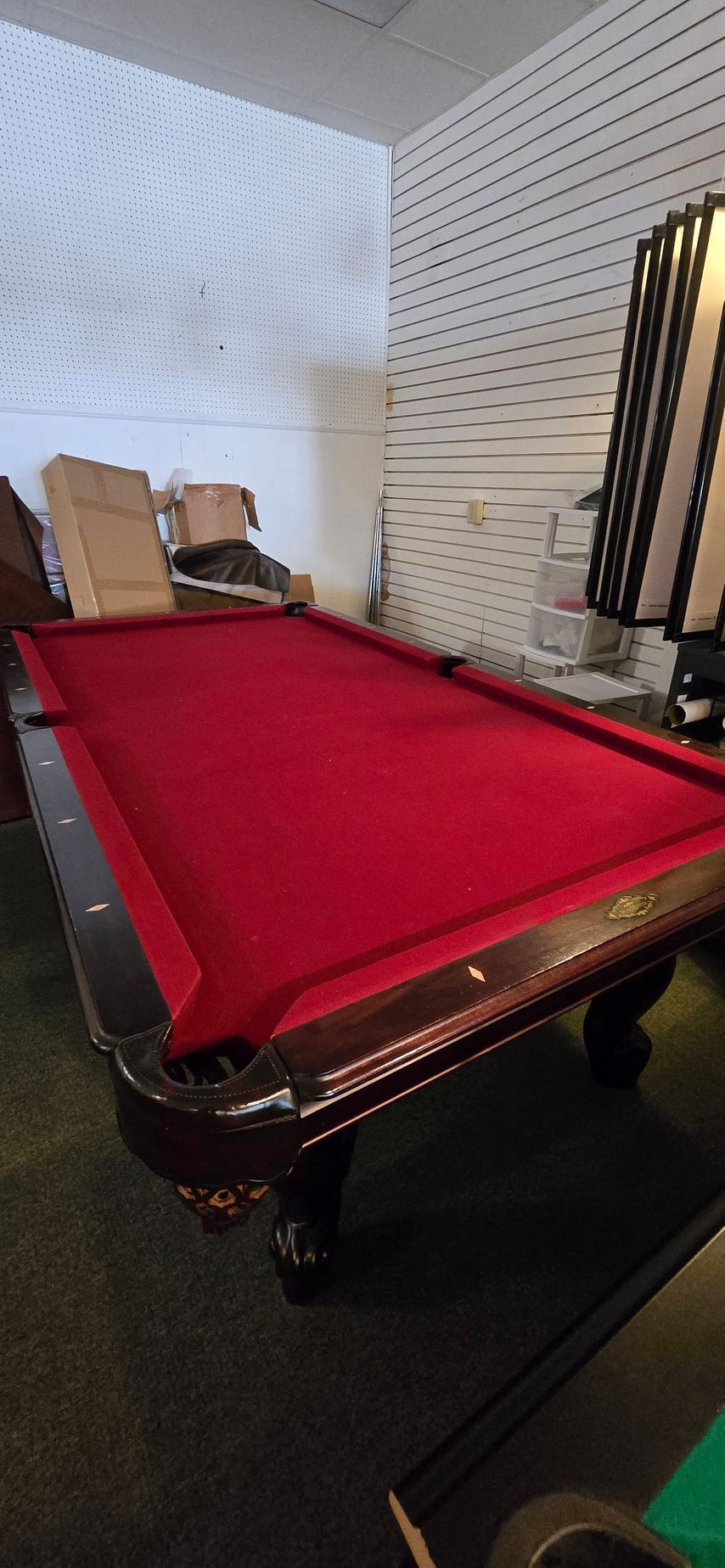 Red-felt pool table in a room with white slatted walls and stacked chairs in the background.