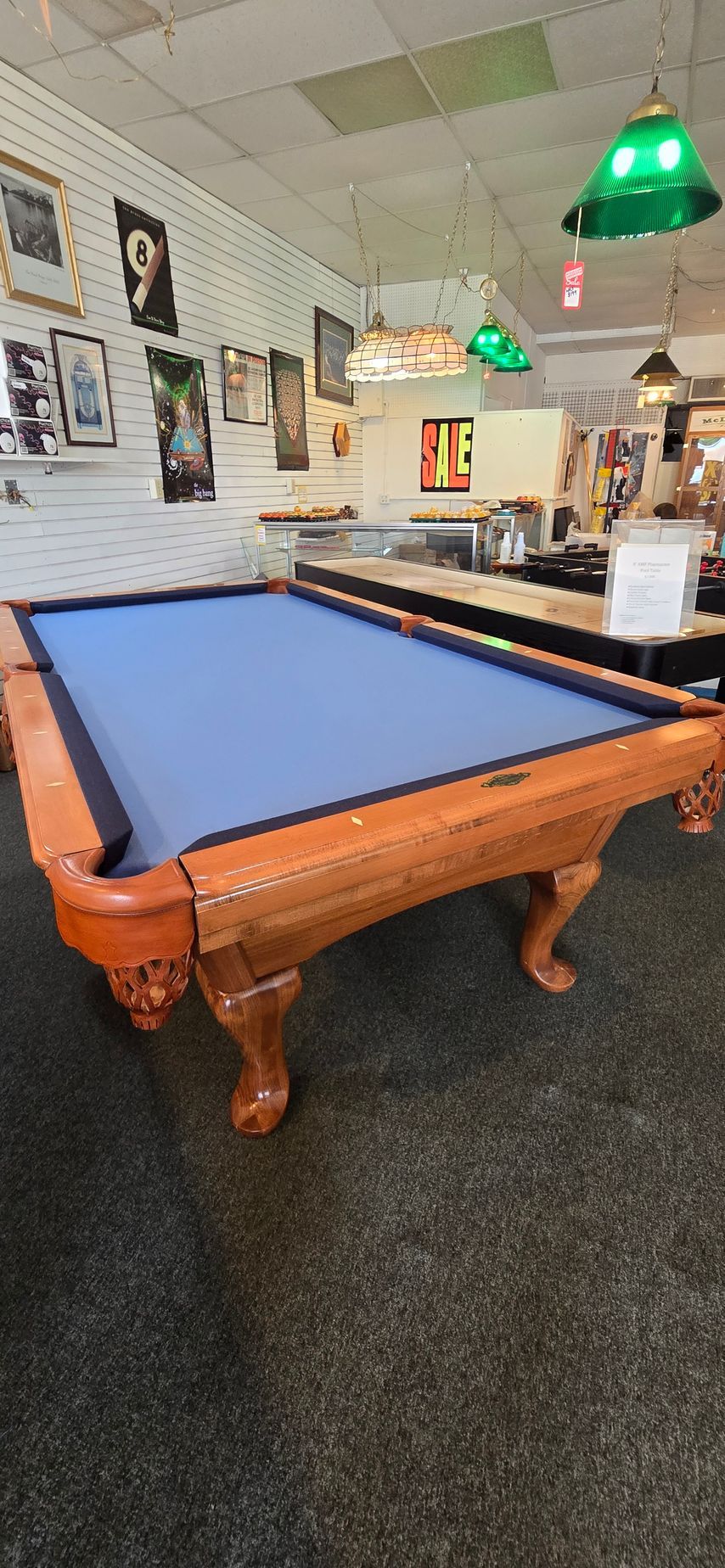 Blue-felt pool table in a game room with posters and green hanging lights.