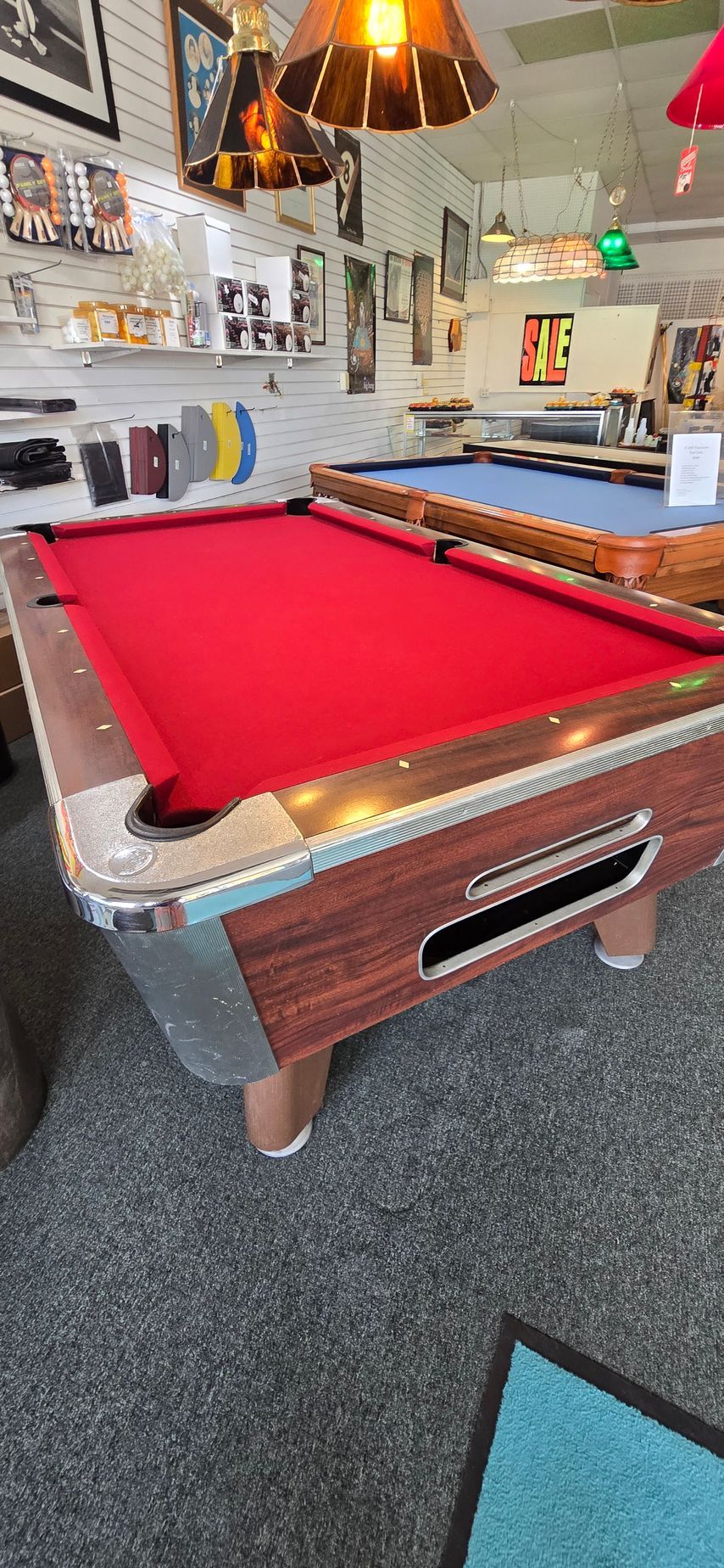 Red felt pool table in a game room with cue racks, posters, and hanging lamps.