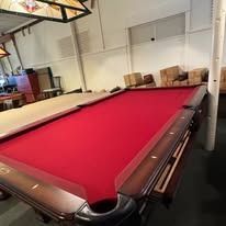 Red-felt pool table in a lounge with chairs and overhead lights, viewed from one corner