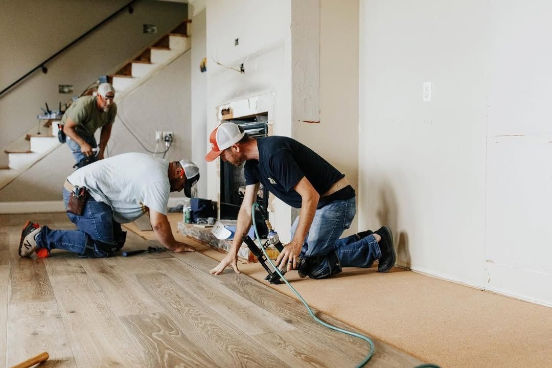 Three construction workers installing wood flooring in a room with stairs and a fireplace.