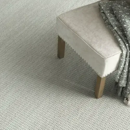 Light-colored rug with a beige ottoman with wooden legs, covered with a gray blanket.
