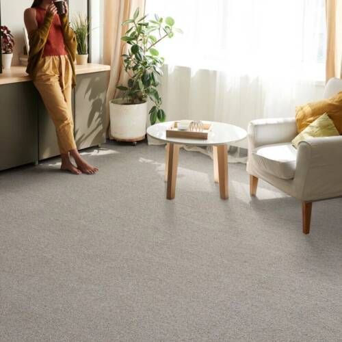 Woman leans on cabinet in a bright room with light gray carpet, chair, coffee table, and large houseplant.