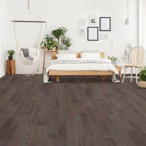 Bedroom with dark wooden flooring, wooden bed, plants, and wall art. White walls and natural light.