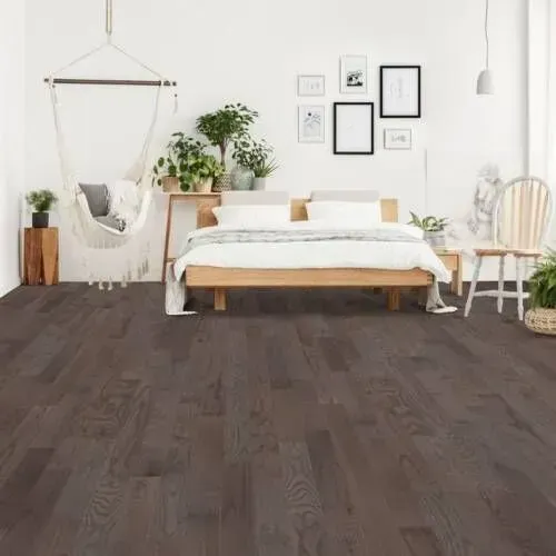 Bedroom with dark wooden flooring, wooden bed, plants, and wall art. White walls and natural light.