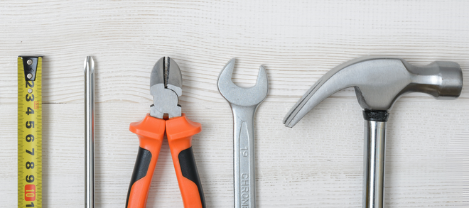 Tools arranged on a light wood surface, including a measuring tape, screwdriver, pliers, wrench, and hammer.