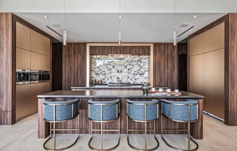 Modern kitchen with wood and gold cabinetry, marble backsplash, and blue upholstered bar stools