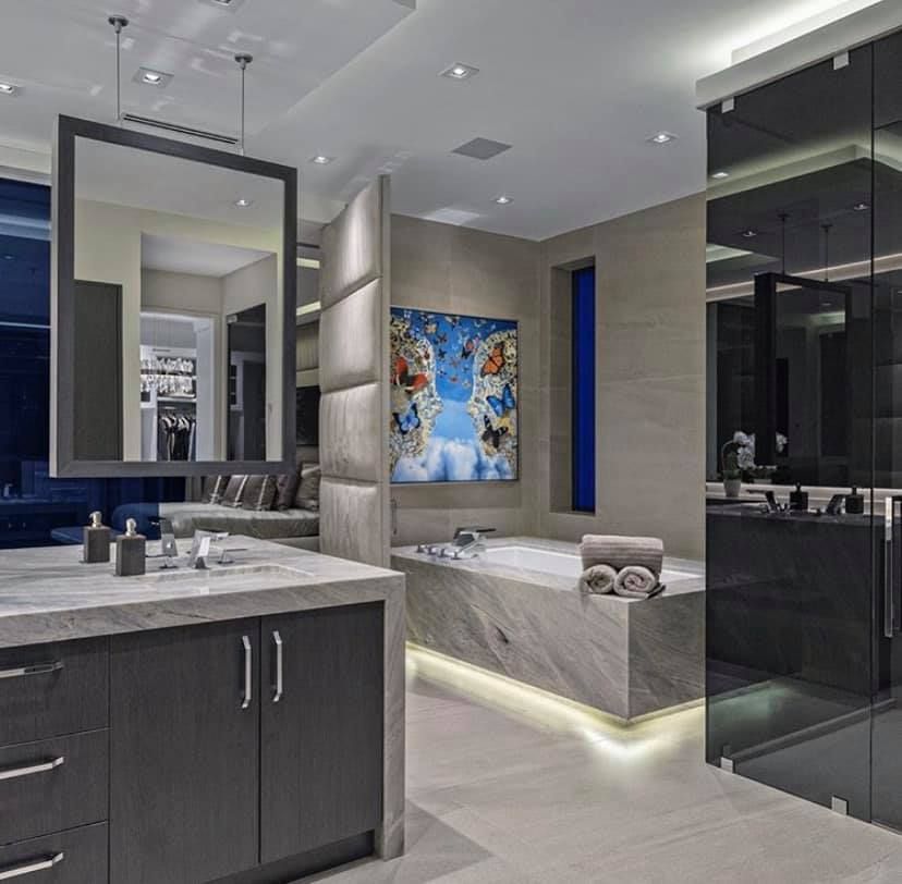 Luxurious modern bathroom with marble, gray cabinets, art, a large mirror, and glass shower