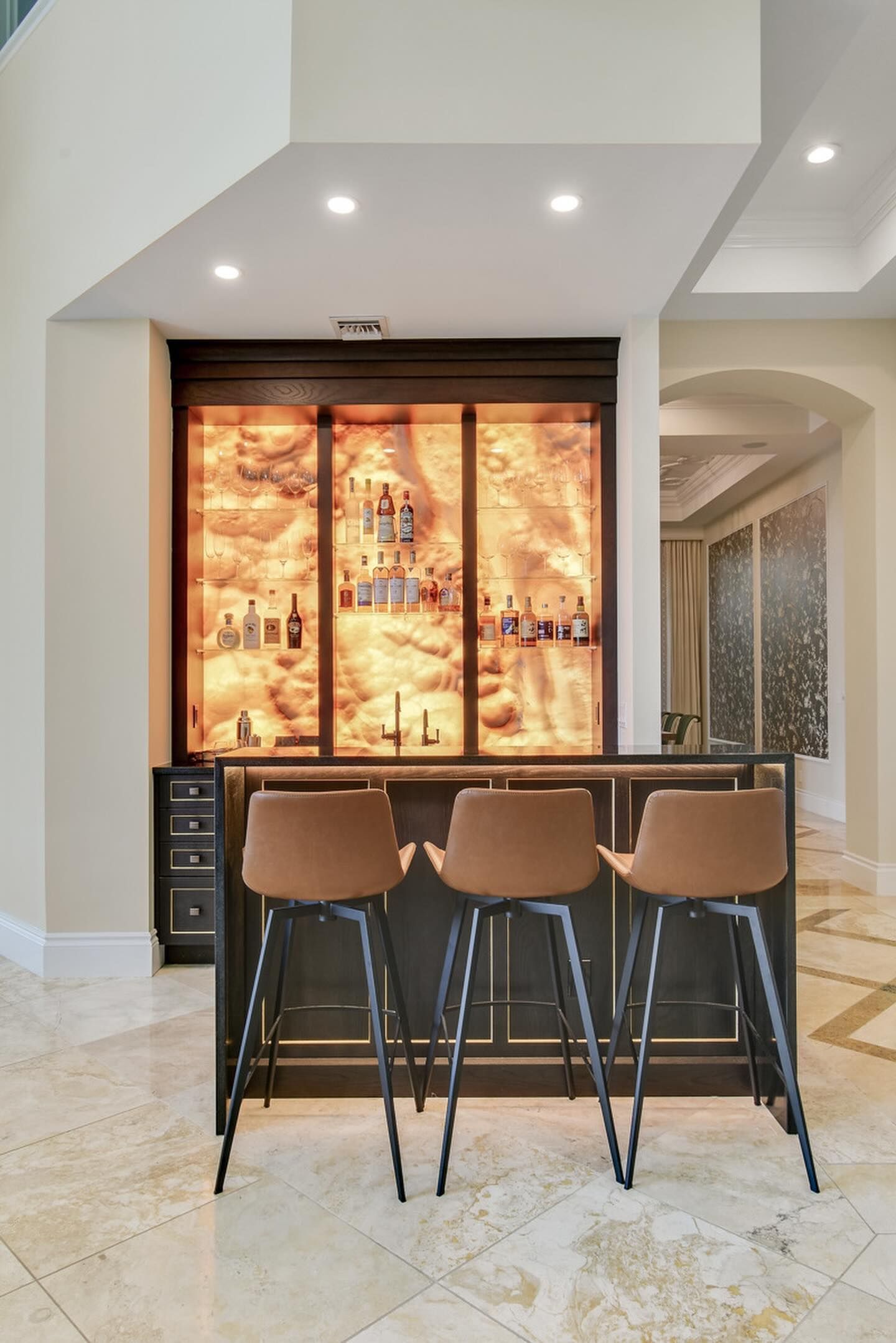 A home bar with three brown stools. Dark cabinets and a lit display of bottles stand behind the bar