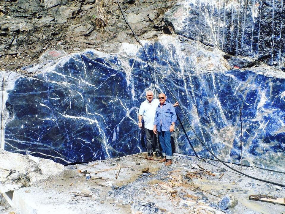 Two men stand before a large slab of blue sodalite stone in a quarry