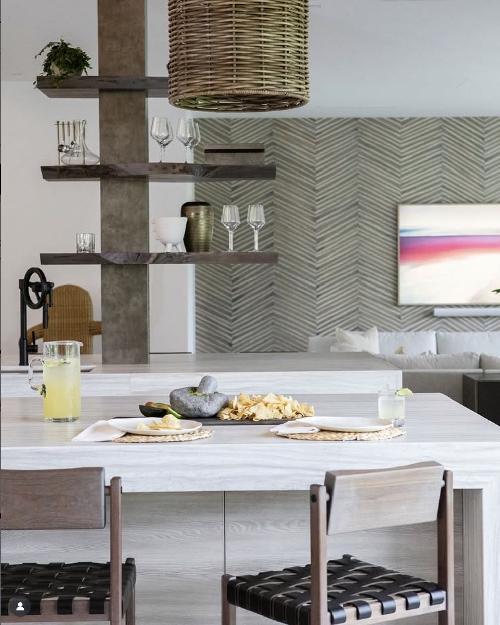 Kitchen with light-colored countertop, bar stools, shelves, woven light fixture, and herringbone wall design