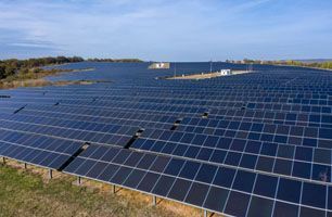 A large solar farm with rows of blue solar panels set in a grassy field under a clear blue sky.