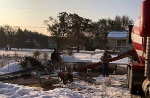 A cement truck pours concrete into a foundation at a snowy construction site as two workers stand nearby.