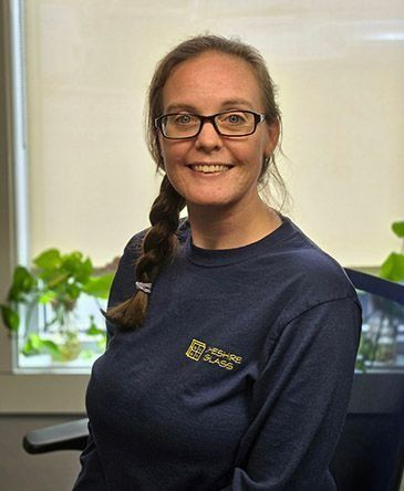 Smiling person with glasses and braided hair in a navy shirt, standing indoors near plants and a window