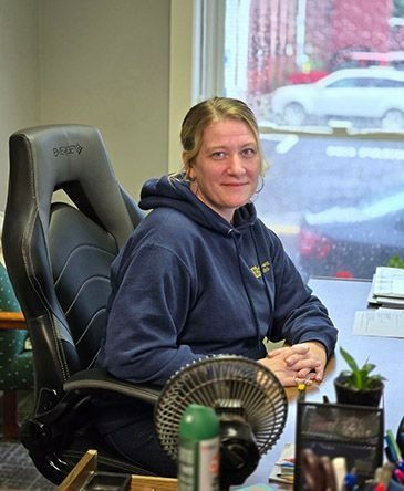 Woman in a navy hoodie seated at a desk, smiling in an office with a window and fan.