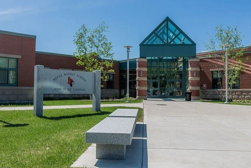 School building entrance with teal glass canopy, sidewalk bench, and sign on a sunny day