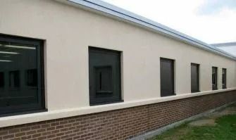 Modern white building with several dark-framed windows and a brick base along a grassy edge