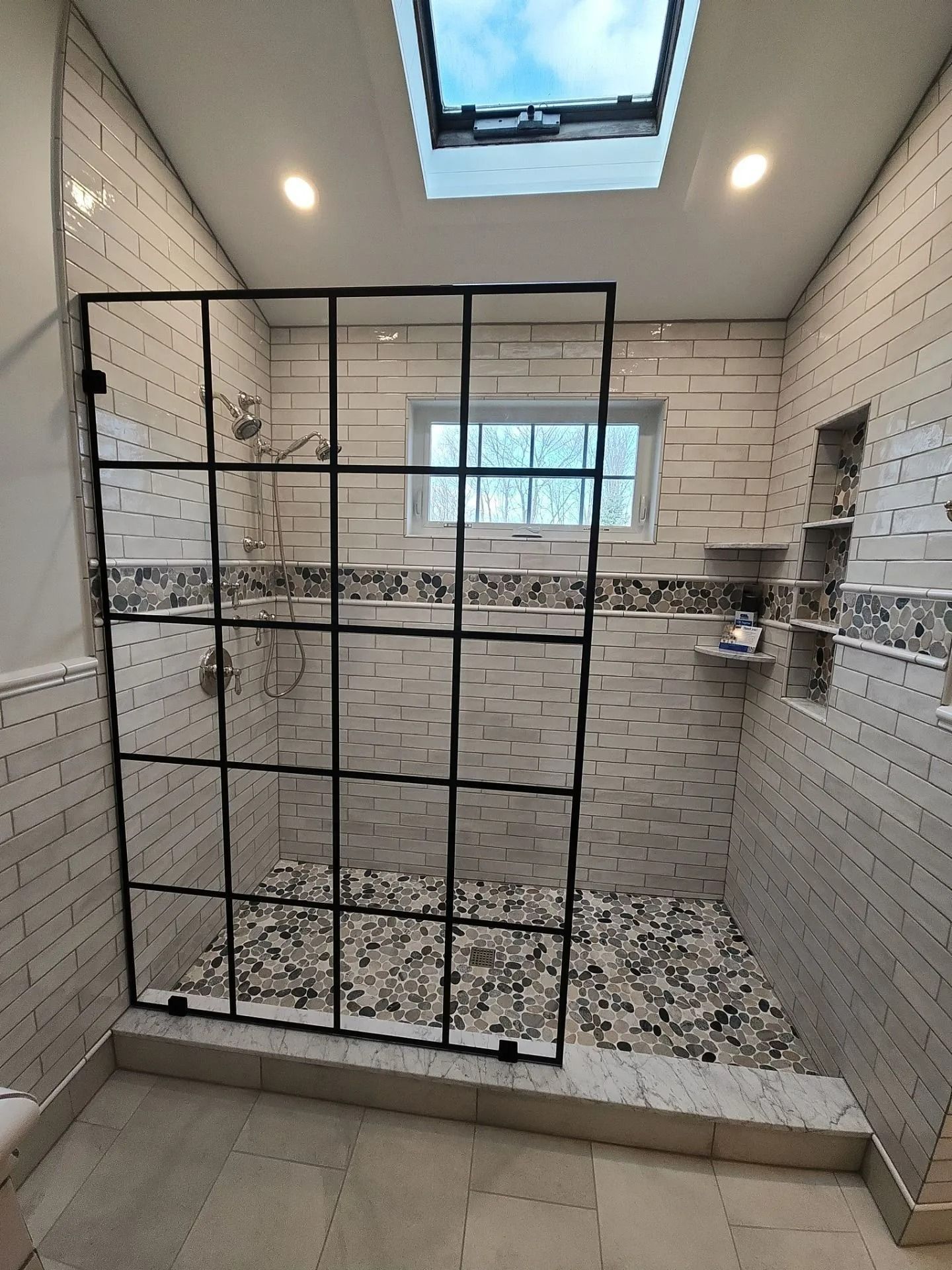 Modern tiled walk-in shower with glass partition, skylight, and patterned pebble floor.