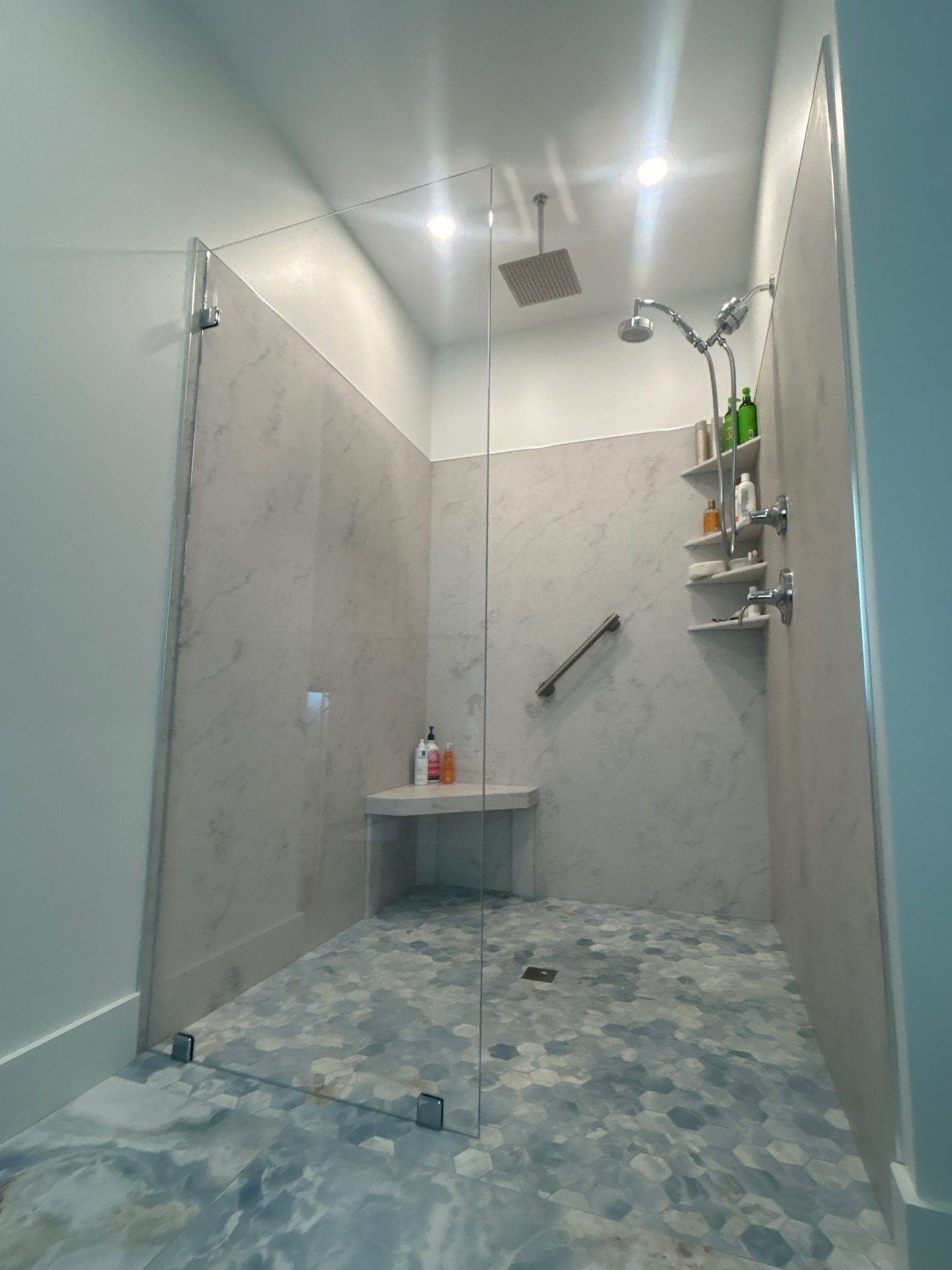 Small modern tiled shower with glass door, rain showerhead, shelf, and blue-patterned floor.