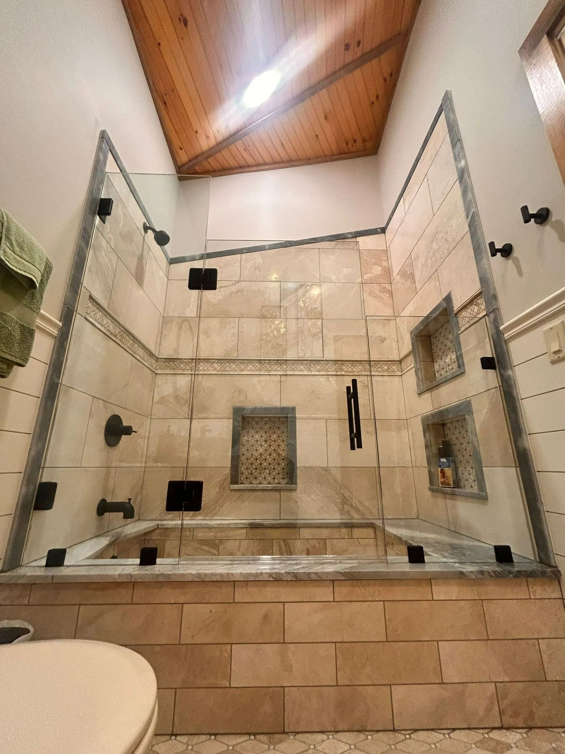 Modern tiled shower with glass doors, beige stone walls, black fixtures, and a wood-paneled ceiling