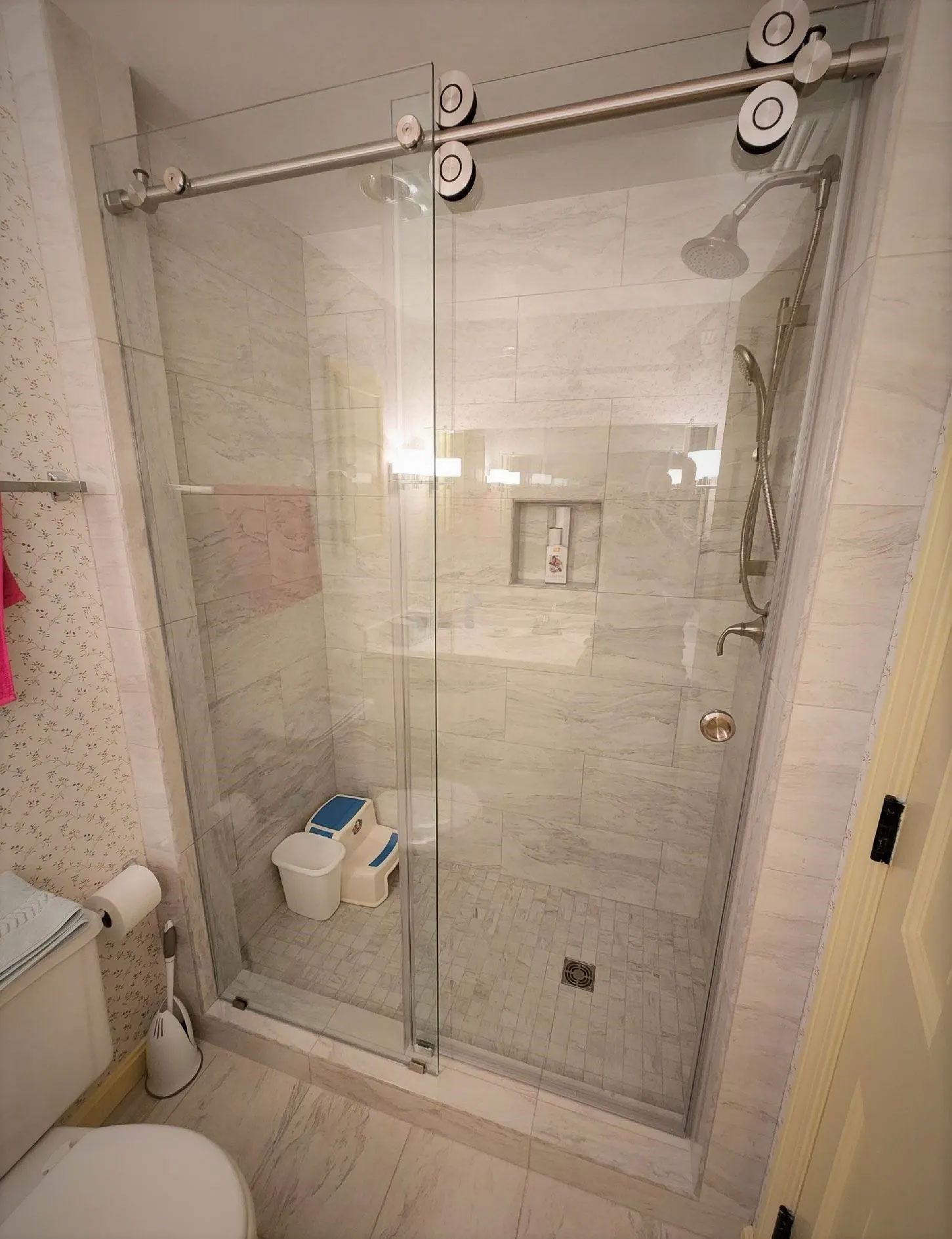 Glass shower enclosure with a sliding door, mounted grab bars, and a shower caddy in a tiled bathroom.