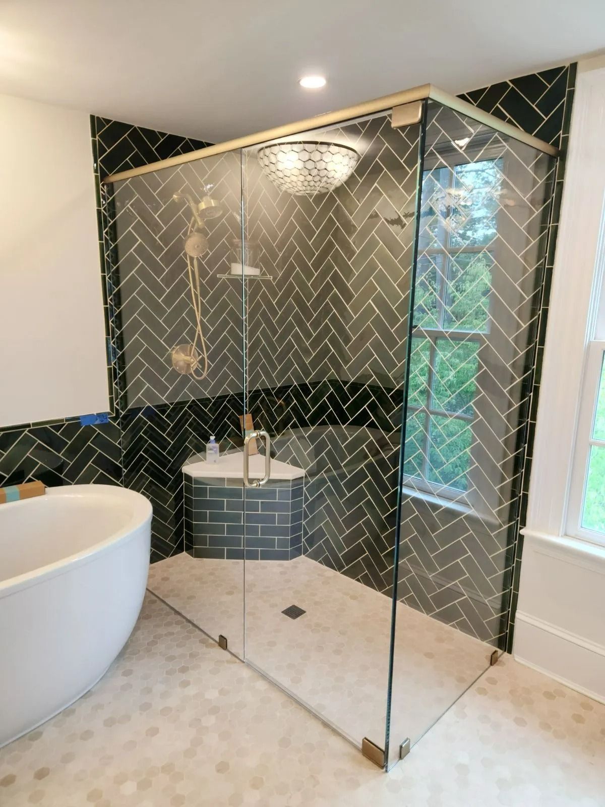 Modern bathroom with black herringbone tile shower, glass enclosure, freestanding tub, and window light