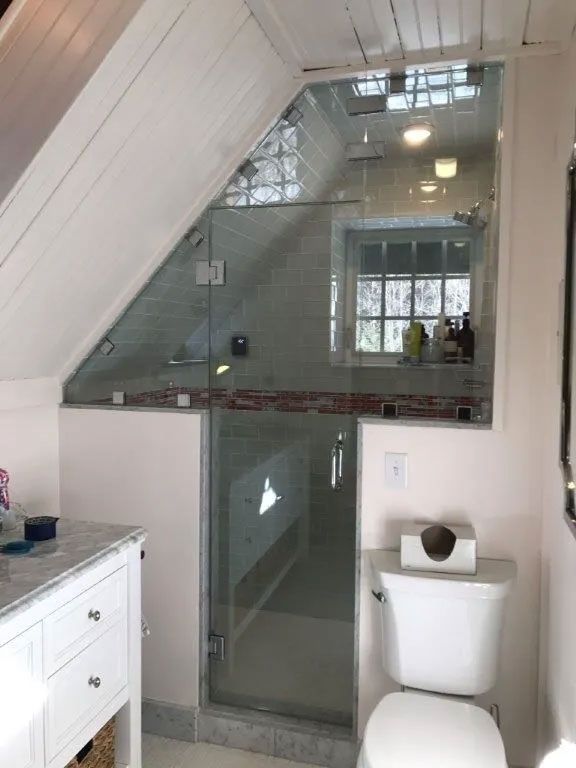 Small bathroom with slanted ceiling, glass shower, and white toilet beside a window.