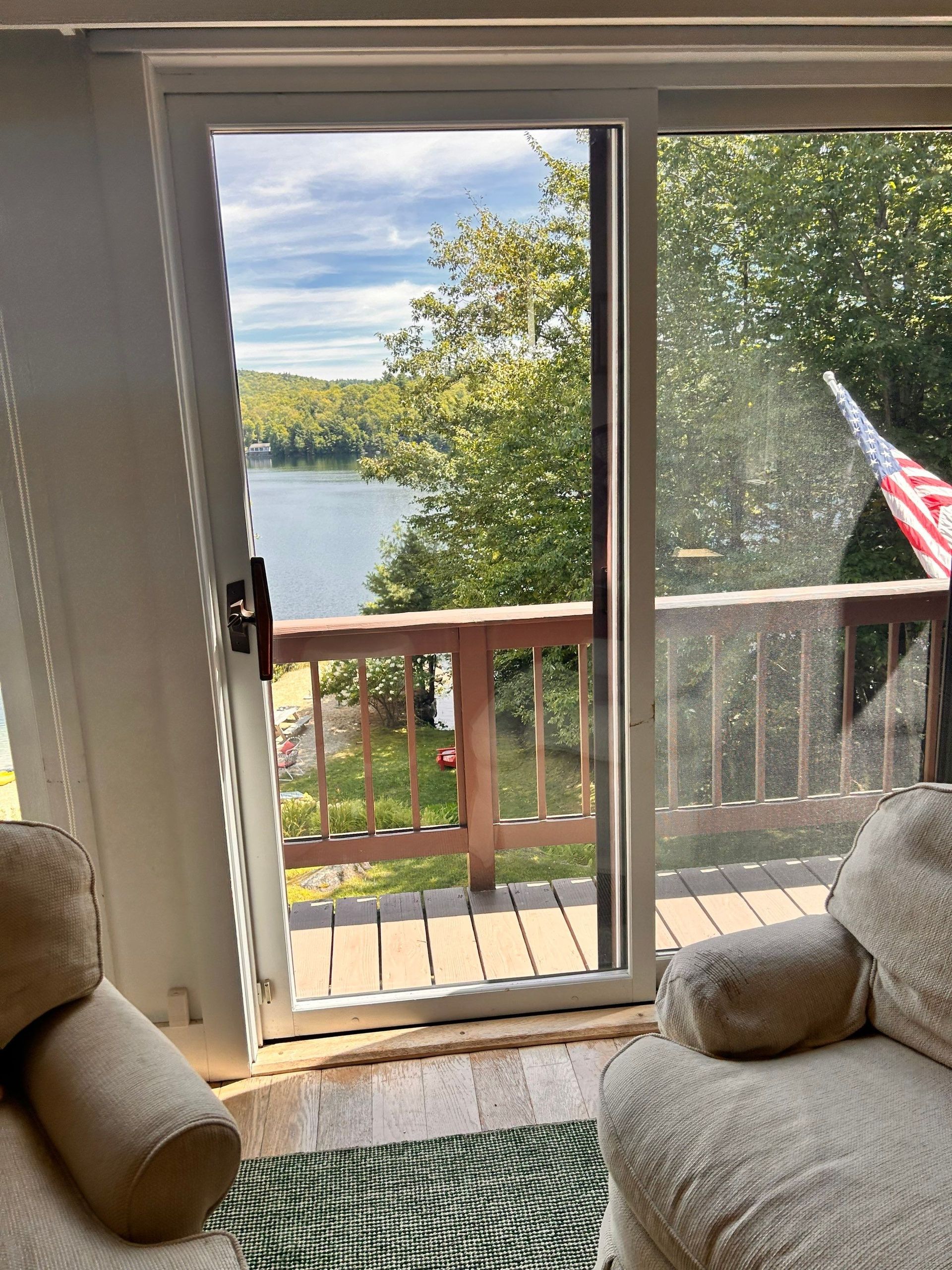 Living room view through open door to a lake and trees, with a wooden deck and patio chair visible