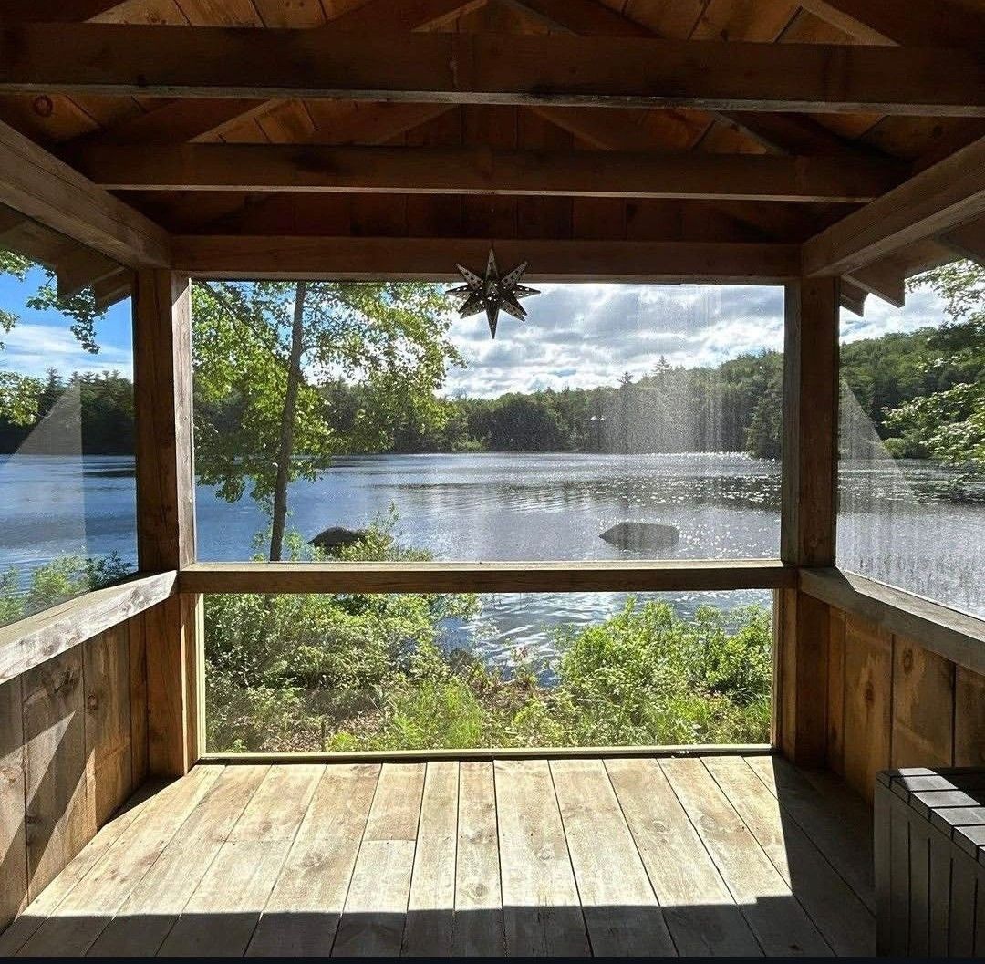 Wooden lakeside gazebo with a bench overlooking a sunlit lake and trees