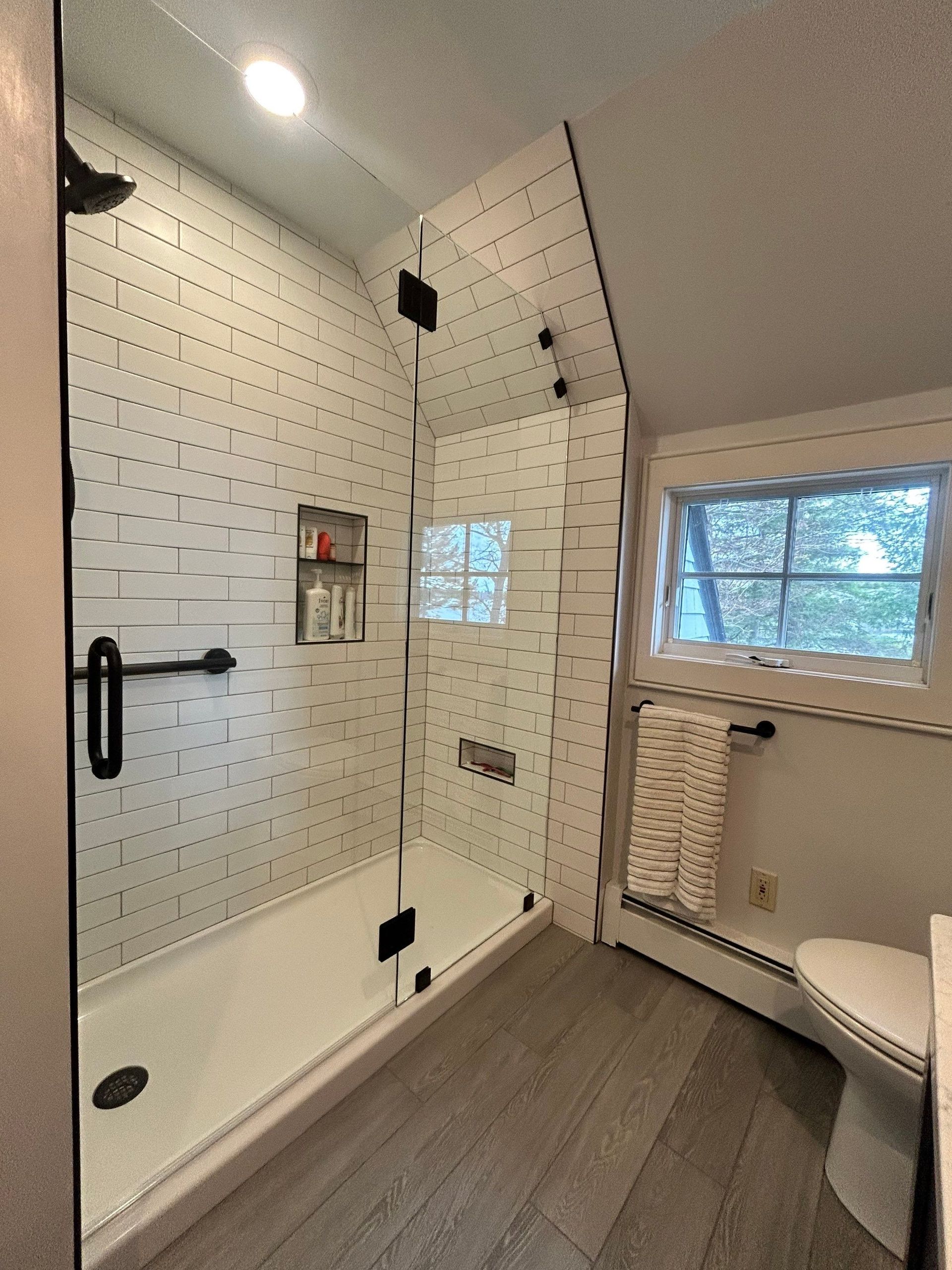 White tiled bathroom with a walk-in shower, black grab bar, and toilet by a small window.