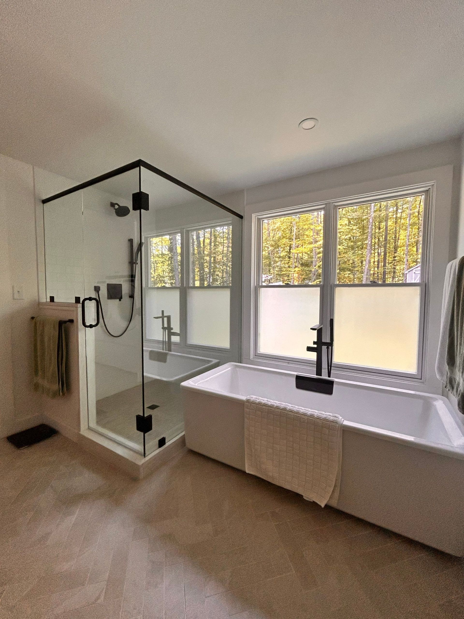 Modern bathroom with glass shower, large tub, and two windows with white blinds