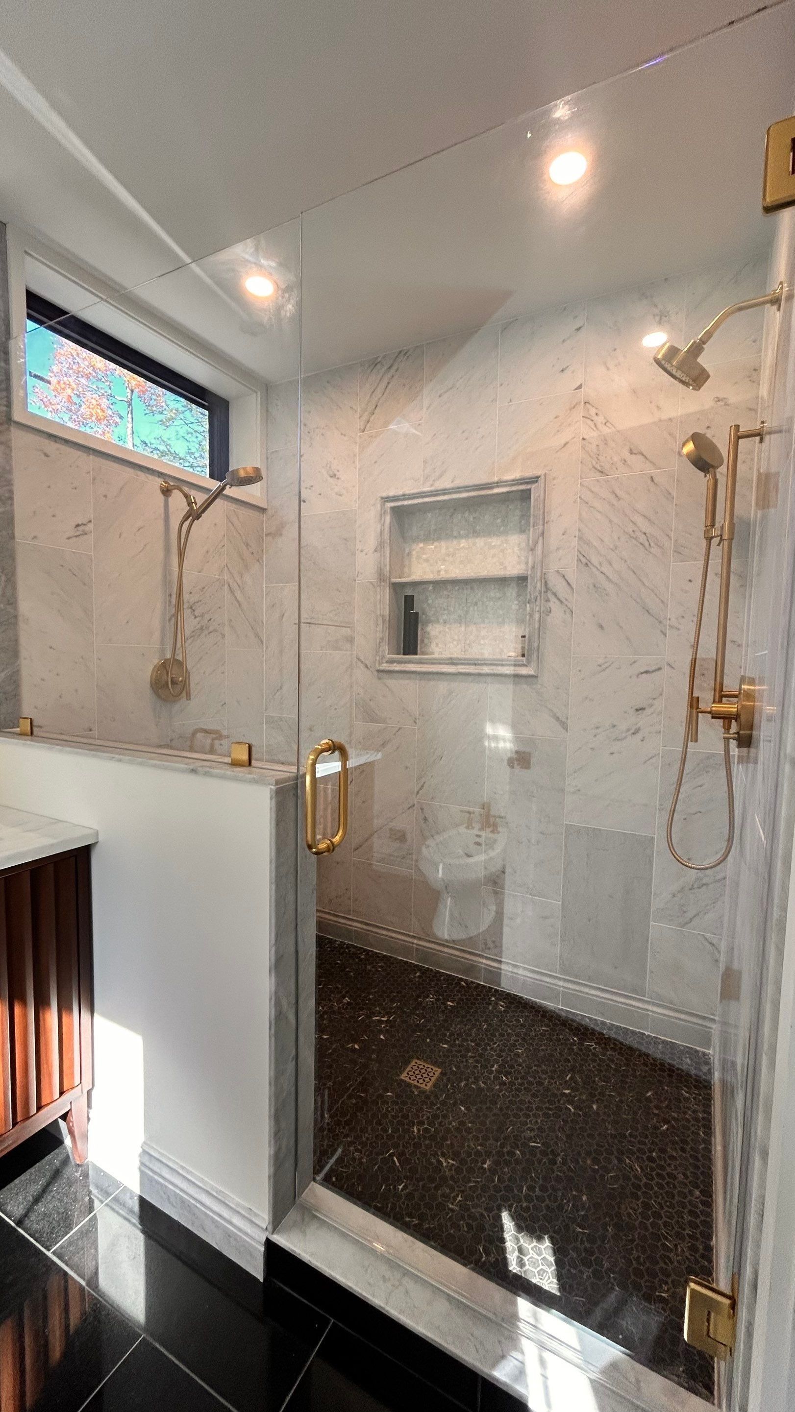 Glass shower enclosure with marble walls, built-in shelf, and brass fixtures in a bright bathroom
