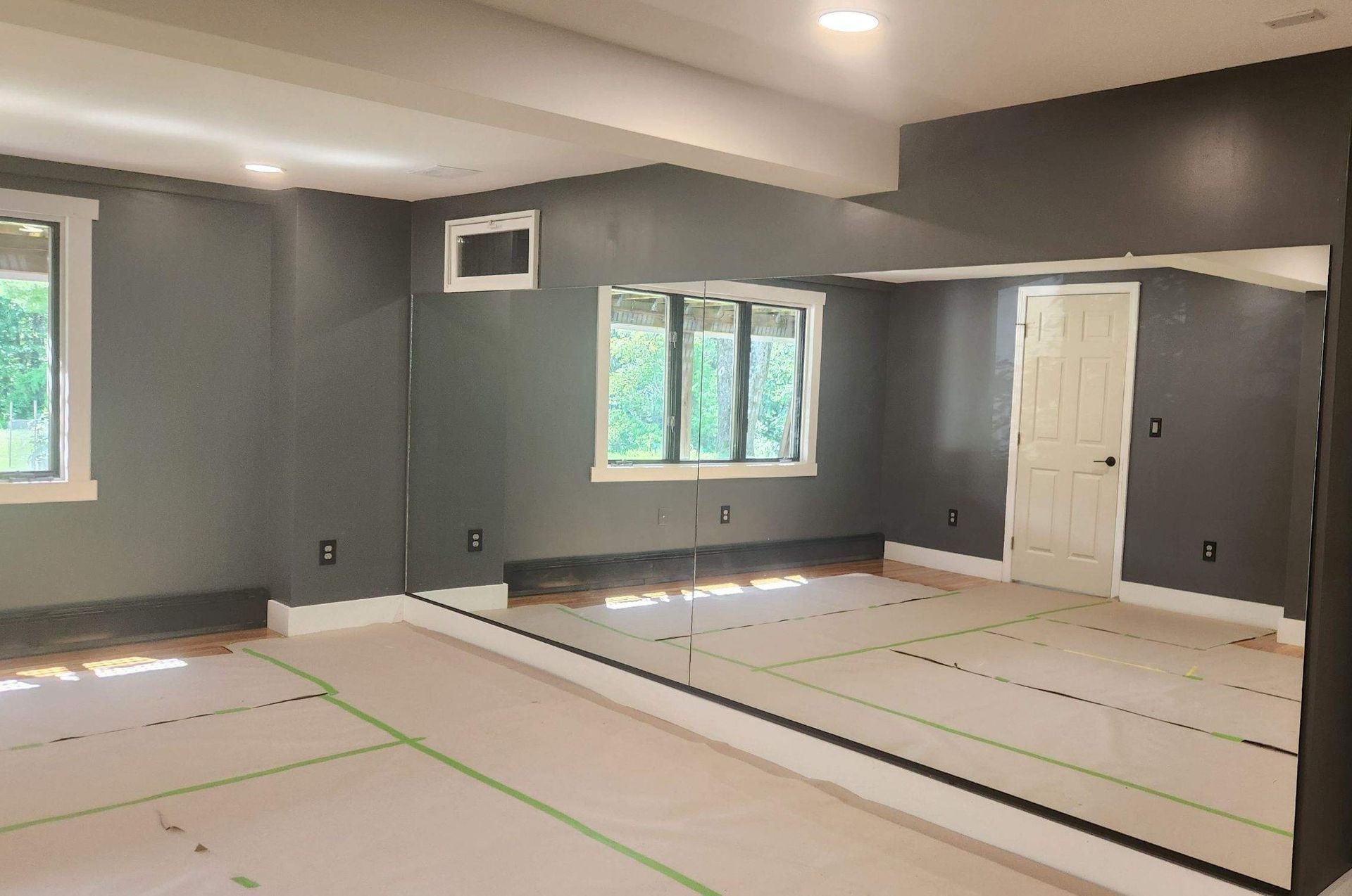 Empty gray room with windows, white door, and taped-off floor covering, likely under renovation