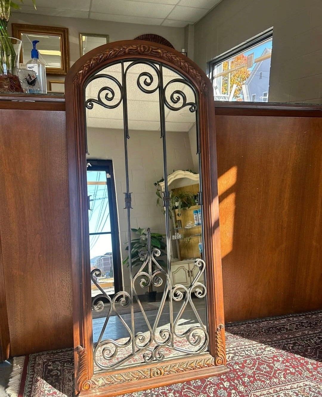 Decorative wrought-iron mirror in a wood-paneled hallway with sunlight and a patterned carpet underneath