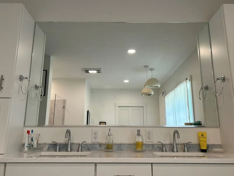 Bathroom vanity with large mirror, double sinks, chrome faucets, and white cabinets under bright ceiling lights
